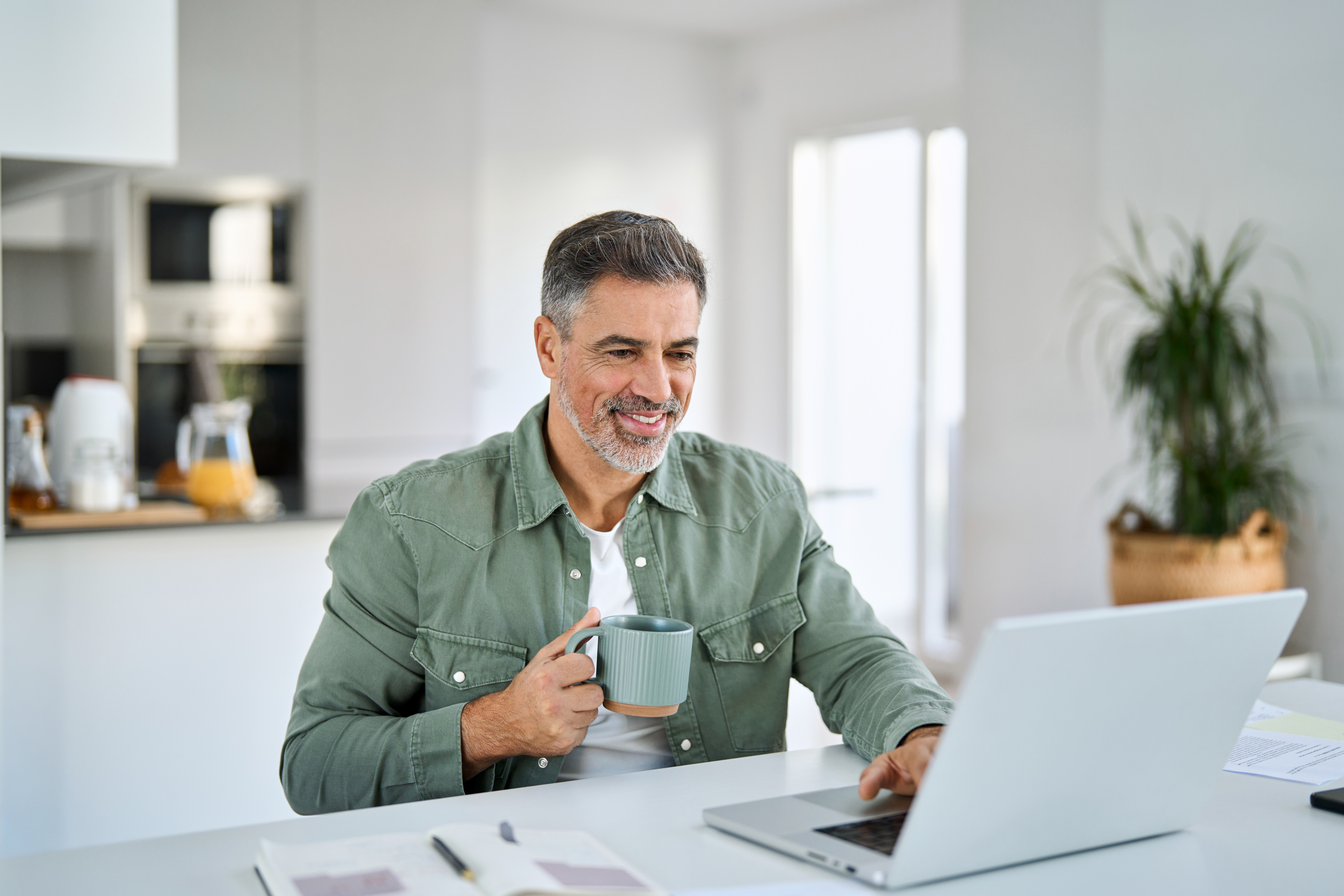 Älterer Mann sitzt mit einer Tasse Kaffee in der Küche am Laptop